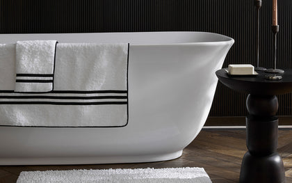 White Newport towel with black striped border draped on a freestanding tub, bath mat below.