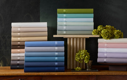 Nocturne sateen sheets stacked in assorted colors on a wood console with hydrangeas.
