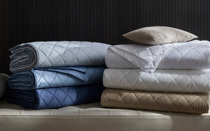 Diamond-quilted Nocturne Quilt bedding stacked in assorted colors on a quilted bench.