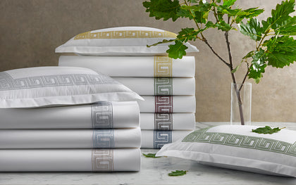 White Ophelia sheets with Greek key embroidered trim in assorted colors stacked on marble with oak leaves.