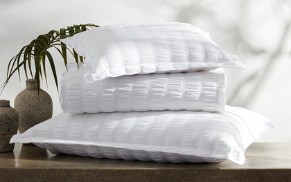 White seersucker-textured Panama bedding stacked on a stone ledge beside a palm frond.