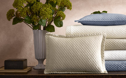 Textured Perla quilted bedding stacked in cream and blue on a console beside hydrangeas.