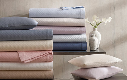 Diamond-quilted Petra bedding stacked in assorted colors on a table beside white ceramic vases.