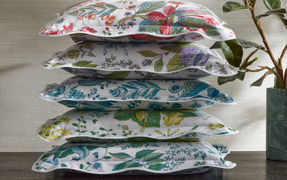 White Pomegranate floral-print pillows in assorted colorways stacked beside a leafy plant.