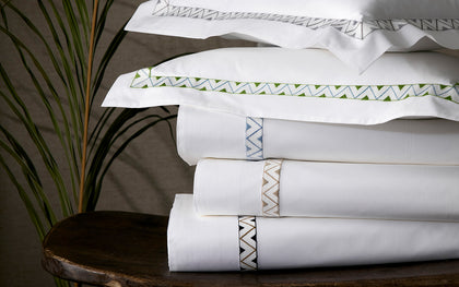 White Prado sheets with zigzag embroidered trim in assorted colors stacked on a wood table.