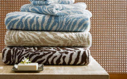 Zebra-print Regine towels in blue, taupe, and brown stacked on a stone ledge with a soap dish.