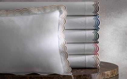 White Stella bedding with scalloped embroidered trim in assorted colors stacked on a marble table.