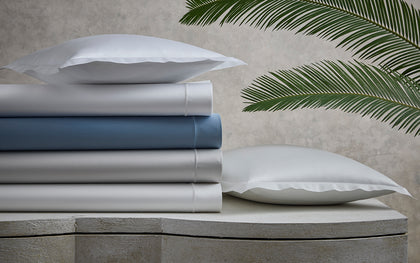 Talita Satin Stitch sheets stacked in white, blue, and silver on a stone bench beside palm fronds.