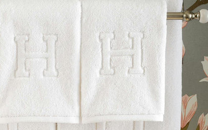 White Auberge towels with a monogrammed letter H in bold relief, hanging on a rod.