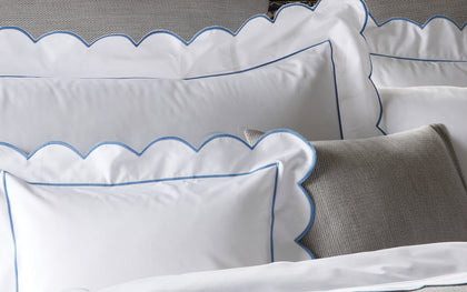 White Butterfield scalloped shams with blue trim, styled on a bed with a gray textured pillow.