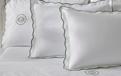 White Camilla Pique scalloped pillows with green trim and monograms, styled on a tufted bed.