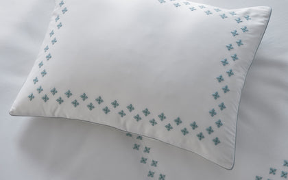 White Casablanca pillow with blue diamond cross-stitch trim, styled on a bed.