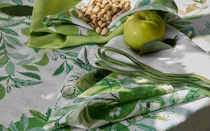 Green Citrus Garden leaf-print tablecloth and napkins set with a green apple and pistachios.