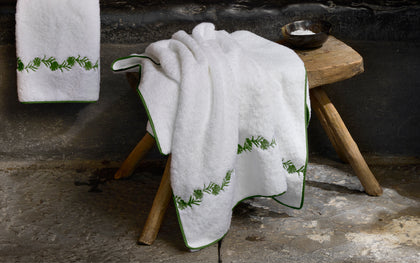 White bath towel with green fern-embroidered trim, draped over a wooden stool against a dark stone wall, with soap and a second towel beside it.
