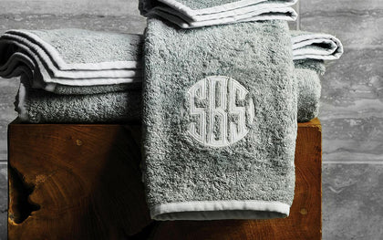 Gray Enzo towels with white trim and a monogram folded on a wooden stool.