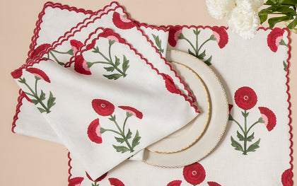 White Gisele placemats and napkins with embroidered red poppies, stacked with a gold rimmed plate.
