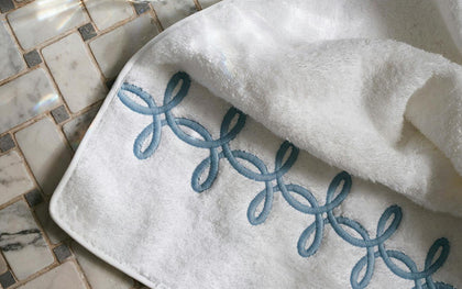 White Gordian Knot towel with blue embroidered knot trim on a marble tile floor.