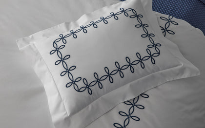 White Gordian Knot pillow with a navy embroidered border on a bed.