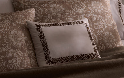 Tan Granada floral print shams layered with a scroll-embroidered pillow on a bed.