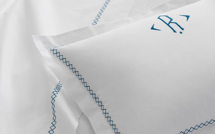 White Hatch monogrammed pillow with blue cross-stitch trim, layered on a bed.