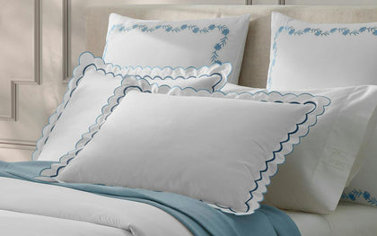 White India scalloped pillows with blue trim, styled on a bed with floral embroidered pillowcases and a blue throw.