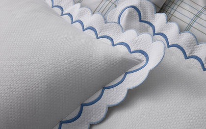 White India Pique scalloped pillow with navy trim, layered on a bed near plaid pillows.