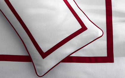 White Louise Pique pillow with a red bordered trim, layered on a bed.