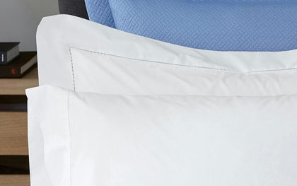 White Luca Hemstitch pillows with a hemstitch trim, layered on a bed with a blue quilted headboard.