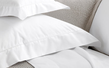 White Milano Hemstitch pillowcases and sheets layered on a bed with a woven headboard.