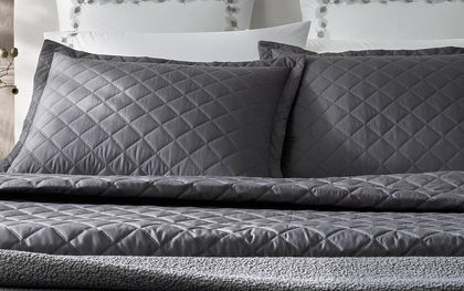Charcoal diamond-quilted Milano Quilt shams and coverlet with vine-embroidered white pillowcases behind.