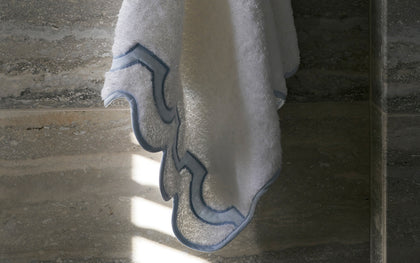 White Mirasol towel with scalloped blue trim hanging beside a glass shower panel.