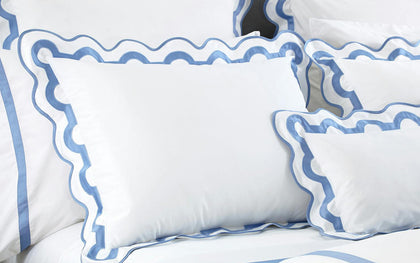 White Mirasol pillow shams with scalloped blue trim layered on a bed.