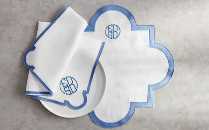 White scalloped Mirasol placemat with blue trim beside monogrammed napkins and a plate.