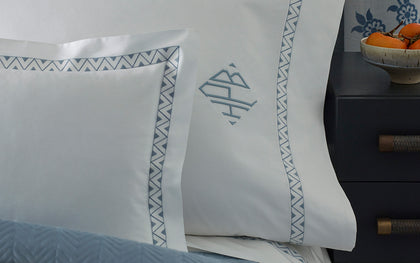 White Prado bedding with blue zigzag embroidered trim and monogram beside a dark nightstand.