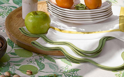 White Scallop Edge napkins with green wavy trim beside stacked plates and fresh fruit.