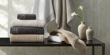 Adelphi towels with greek-key trim in white, tan, and charcoal, hung and stacked beside calla lilies.