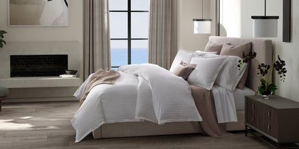 White Amalfi pinstripe bedding styled on a bed with a taupe throw near an ocean-view window.