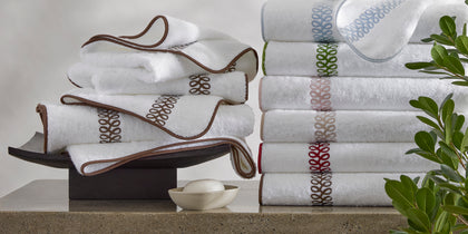 White Astor Braid towels with loop-embroidered trim in assorted colors, stacked beside soap and greenery.