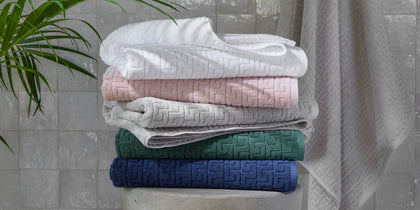 Athena greek-key textured towels in white, pink, gray, green, and navy, stacked on a round stool.