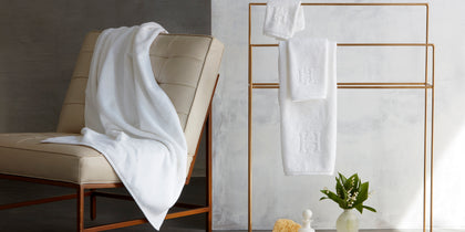 White Auberge monogrammed towels on a gold towel rack, beside a soap tray and flowers.
