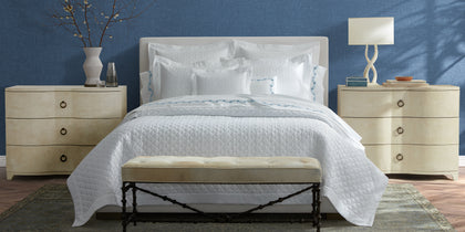 White Ava quilted bedding with blue floral embroidered trim, styled on a bed against a blue wall.