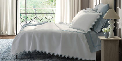 White Aziza Matelasse quilted bedding with blue wavy trim, styled on a bed near a balcony window.