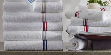 Stacked white Bel Tempo towels with colorful striped trim, soap, and a leafy plant.