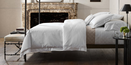 White Bergamo Hemstitch bedding with a tan quilted throw, styled on a bed near a marble fireplace.