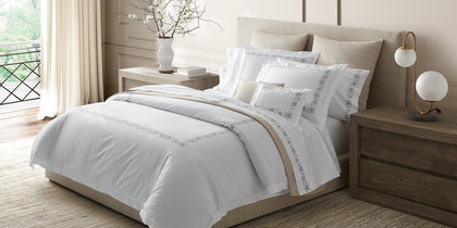 White Callista bedding with gray floral embroidered trim, styled on a bed near a window.