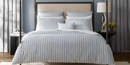 Blue and white striped Calypso bedding styled on a bed with an upholstered headboard.