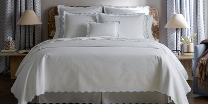 White Camilla Pique scalloped bedding with light blue trim, styled on a bed near a floral headboard.