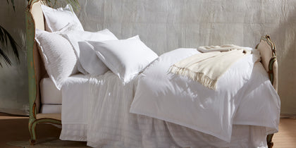 White Ceylon Satin Stitch bedding with a tan fringed throw, styled on a bed with an ornate headboard.
