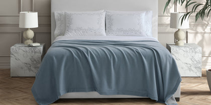 Slate blue Cosmo blanket draped on a bed with zigzag-embroidered shams.