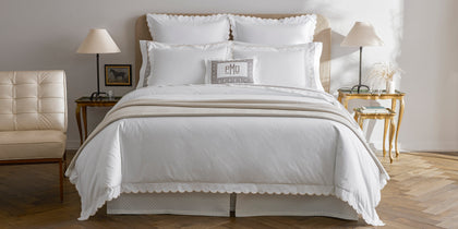 White Dakota scalloped bedding with tan greek-key trim, styled on a bed with a monogrammed pillow.